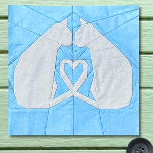 May include: A fabric art piece featuring two white cat silhouettes facing each other, forming a heart shape with their tails. The background is a light blue geometric pattern, creating a patchwork design.