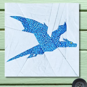 May include: A patchwork design featuring a blue dragon with a leopard print pattern against a white background. The dragon has outstretched wings and an orange eye. The artwork is made of fabric.