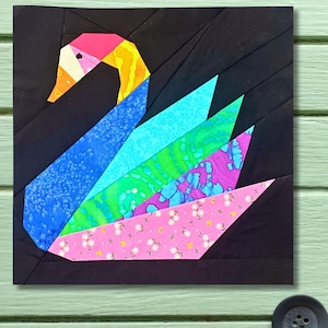 May include: A colorful, geometric swan quilt block. The swan features a black background with a pink, yellow, and orange head, and blue, green, and pink patterned feathers. The quilt block is square and set against a light green background.
