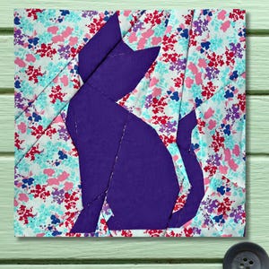 May include: A square fabric art piece featuring a purple cat silhouette against a floral patterned background. The cat is made of solid purple fabric, while the background is a patchwork of floral fabric in shades of pink, blue, and red.