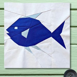 May include: A geometric design of a blue fish on a white background. The fish is constructed from various shades of blue fabric, with a white eye and fins. The background is a patchwork of white fabric.