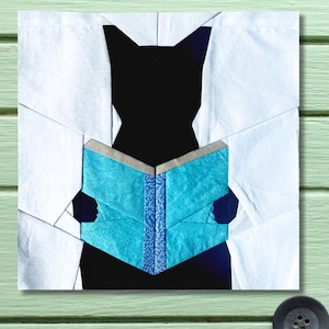 May include: A fabric artwork depicting a black cat silhouette absorbed in a book. The open book has blue patterned pages and a grey spine. The cat is set against a white background, providing a strong contrast.
