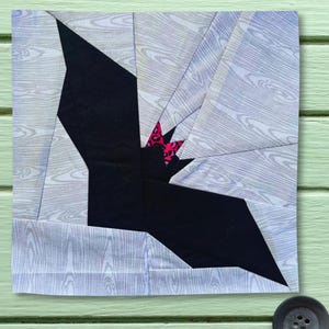 Puede incluir: Un bloque de tela cuadrado para acolchar que presenta la silueta central de un gato negro con un pequeño lazo de estampado de leopardo rosa y negro. El fondo consta de segmentos de tela unidos con un patrón de vetas de madera gris claro.