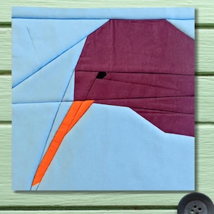 May include: A geometric fabric art piece featuring a bird design. The bird's head is a deep burgundy color, with an orange beak and a small black eye. The background is light blue, and the artwork is mounted on a light green surface.