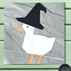 May include: A geometric quilt block featuring a white goose wearing a black witch's hat. The goose has an orange beak and feet. The background is gray, with a textured, angled pattern.