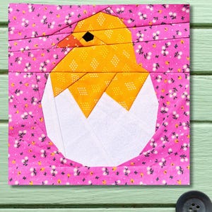 May include: A fabric square with a patchwork design of a yellow chick hatching from a white egg. The chick has a black eye and orange beak. The background is pink with a floral pattern.
