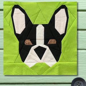 May include: A fabric art piece featuring a geometric design of a French Bulldog's face. The dog's face is black and white with brown accents for the eyes, set against a green background. The design is square and appears to be a quilt block.