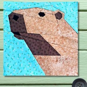 Peut inclure: Une œuvre d'art textile carrée représentant un visage d'ours au design géométrique. Le visage de l'ours est composé de différents motifs de tissu, notamment des imprimés floraux bruns et beiges, et un fond floral bleu clair.