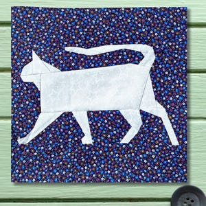 Puede incluir: Un bloque de tela cuadrado con una silueta de gato blanco sobre un fondo floral azul oscuro. El gato está representado caminando. Este bloque es probablemente parte de un proyecto de acolchado o textil más grande.