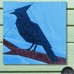 May include: A bluebird silhouette perched on a branch, crafted from fabric. The bird is a dark blue with a speckled pattern, set against a light blue background with a polka dot design. The branch is brown with a floral pattern.