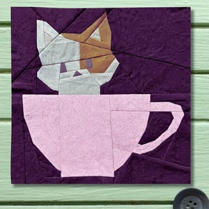 May include: A fabric art piece featuring a geometric design of a cat in a teacup. The cat's head is white and brown, set against a deep purple background. The teacup is light pink.