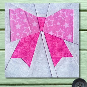 Può includere: Un blocco di tessuto quadrato con un disegno a fiocco. Il centro e le code del fiocco sono rosa pieno, mentre i passanti del fiocco hanno un motivo a stelle rosa su sfondo bianco. Lo sfondo è grigio chiaro.