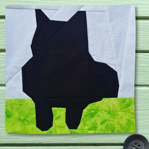 May include: A square fabric quilt block featuring a solid black silhouette of a cat standing in profile. The background consists of light gray fabric with a subtle white floral pattern, while the foreground is bright green mottled fabric.