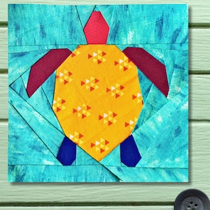 May include: A colorful fabric quilt block featuring a turtle design. The turtle has a yellow patterned shell, red head, burgundy flippers, and blue feet. The background is a turquoise blue with a textured appearance.