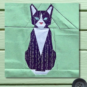 May include: A geometric patchwork cat design in shades of purple, white, and mint green. The cat has white ears, a white chest, and turquoise eyes. The cat is sitting on a mint green background.