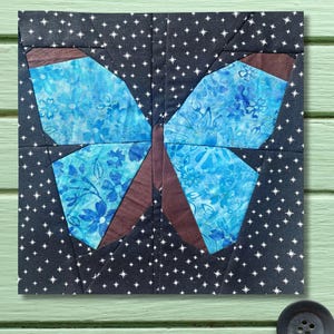 May include: A square fabric block featuring a butterfly design. The butterfly's wings are a vibrant blue with floral patterns, outlined in brown. The background is dark blue with white star-like shapes. The butterfly's body is brown.