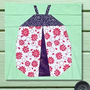 May include: A fabric block featuring a ladybug design. The ladybug's body is white with red floral patterns, and the head is navy blue with a swirl design. The background is light green.