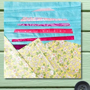 May include: A colorful patchwork quilt square featuring a landscape design. The sky is light blue, with pink and red horizontal stripes representing the horizon. The foreground features a yellow floral pattern, creating a vibrant and artistic textile piece.