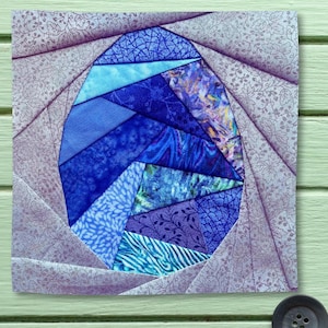Op de afbeelding: Een vierkant stoffen quiltblok met een centrale ovale eivorm, opgebouwd uit diverse blauwe en paarse stoffen stroken met patronen. De omliggende rand bestaat uit lichtpaarse stoffen stroken met een subtiel bloemmotief.