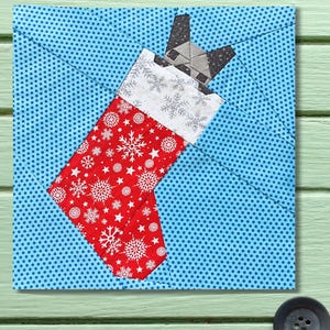 Peut inclure: Une chaussette de Noël en tissu avec un motif de flocons de neige rouges, un revers blanc et une tête de chat grise qui dépasse. La chaussette est posée sur un fond bleu à pois.