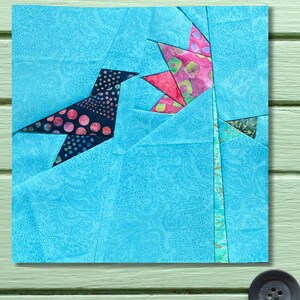 Puede incluir: Un bloque de tela cuadrado con un diseño de patchwork de un pájaro y una flor. El pájaro es azul oscuro con lunares rosas, y la flor es rosa y verde. El fondo es un estampado de cachemira azul claro.