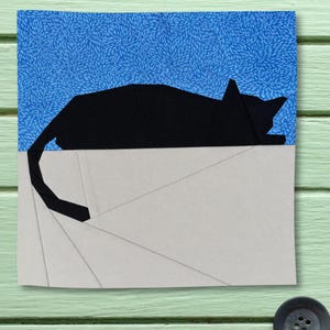 Peut inclure: Un bloc de courtepointe carré présentant la silhouette d'un chat noir appliquée sur un fond bleu à motif floral blanc. La partie inférieure se compose de segments de tissu gris clair disposés en motif géométrique avec des coutures visibles.