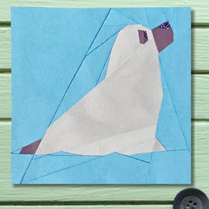 May include: Origami artwork of a seal in white paper, set against a light blue backdrop. The seal has a purple accent on its head and flipper. The background is a patchwork of blue paper, creating a geometric design.