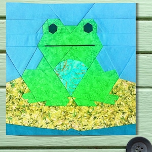 May include: A fabric block with a geometric frog design. The frog is green with a patterned belly, set against a blue and yellow background. The frog's eyes are black, and the design is reminiscent of a quilt block, suitable for crafting projects.