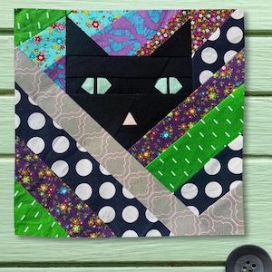 May include: A square fabric quilt block featuring a central black cat face with light green eyes and a small pink nose. The background is composed of diagonal strips of fabric in various patterns, including white polka dots, floral prints, and green.