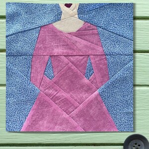 May include: A fabric block featuring a geometric design of a dress. The dress is primarily pink with blue accents and a floral pattern. The neckline is a light beige color. The block is square and appears to be part of a quilt.