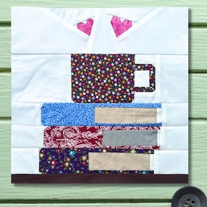 May include: A patchwork wall hanging depicting a coffee cup and a stack of books. The cup has a floral design, and the books have patterned covers in blue, red, and purple. The background is white with pink heart shapes.