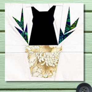 May include: A fabric art piece featuring a black cat silhouette in a floral pot with green and blue leaves. The pot has a gold and white floral pattern, set against a white background.