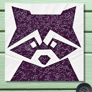 May include: A square fabric block featuring a geometric design of a raccoon's face. The raccoon's face is white and purple, with a textured pattern in the purple areas. The block is set against a light green wooden background.