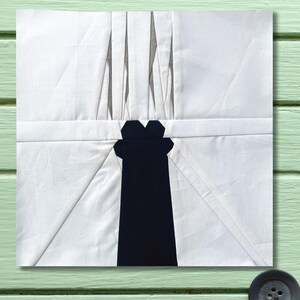 Könnte beinhalten: Ein Stoffkunstwerk in Weiß und Marineblau. Das Design zeigt eine zentrale marineblaue Form mit einem stilisierten Oberteil und weiße Stoffabschnitte mit Falten und schrägen Linien. Das Stück ist quadratisch und liegt auf einer hellgrünen Holzoberfläche.