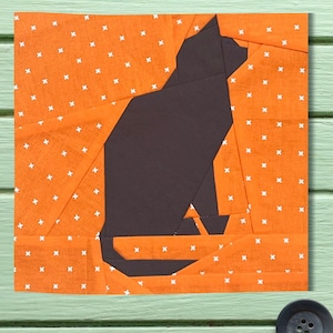 May include: A black cat silhouette paper pieced quilt block on an orange fabric with white x's.