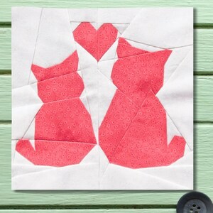 May include: A fabric patchwork square featuring two stylized red cats facing each other, with a red heart between them. The cats and heart are made of patterned fabric, set against a white background.