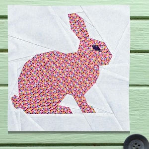 Könnte beinhalten: Ein farbenfrohes Stoffhasen-Design im Patchwork-Stil mit rosa, gelben und blauen herzförmigen Mustern. Der Hase ist vor einem weißen Hintergrund mit geometrischen Linien platziert, was eine moderne, handgefertigte Ästhetik erzeugt. Das Design ist wahrscheinlich für ein Quilt- oder Stoffhandwerksprojekt.