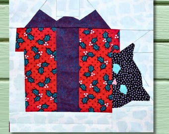 Op de afbeelding: Een patchwork stofblok met een rode geschenkdoos met hulstbessenpatroon en een paarse strik. Een zwart-witte kat is gedeeltelijk zichtbaar. De achtergrond is lichtblauw met een sneeuwvlokkenpatroon.