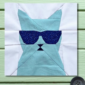 May include: A square fabric block depicting a cat's face with sunglasses. The cat is light blue, with dark blue sunglasses and a small dark blue bow tie. White fabric frames the cat's face.