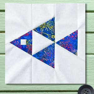Puede incluir: Un bloque de edredón cuadrado con un diseño de pez. El pez está compuesto de papel blanco y triángulos azules con un estampado floral. Un pequeño cuadrado blanco forma el ojo. El fondo es verde claro.