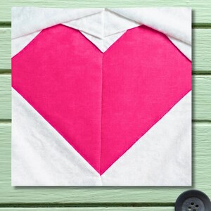 May include: A square fabric block featuring a bright pink heart shape against a white background. The heart is centered and takes up most of the space, with the white fabric forming a border around it. The block is likely part of a larger quilt or craft project.