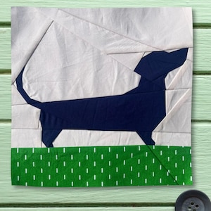 May include: A fabric art piece featuring a navy blue dachshund silhouette against a white and light gray background. The dog stands on a green strip with white vertical dashes, resembling grass. The artwork is square and appears to be a quilt block.