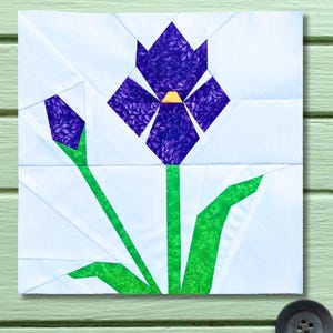 May include: A square fabric block featuring a geometric design of a purple iris flower with a yellow center, green stem and leaves, and a purple bud. The flower is set against a white background.