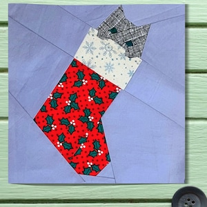 May include: A fabric Christmas stocking with a cat design. The stocking is red with a holly berry pattern. The cat's head is gray and white, and the body is white with snowflakes. The background is light blue.