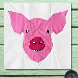 Peut inclure: Un bloc de tissu rose et blanc représentant un visage de cochon géométrique. Le cochon a de grandes oreilles roses, un museau rose avec des détails à pois noirs et blancs et deux yeux à pois noirs et blancs. Le fond est blanc.
