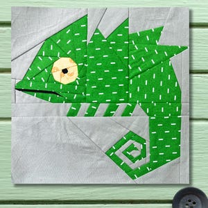 Puede incluir: Un bloque de tela con un diseño de iguana verde. La iguana está hecha de tela verde con guiones blancos, sobre un fondo gris. El ojo de la iguana es un círculo beige con un centro negro. El diseño es cuadrado.