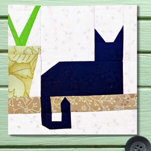 May include: A fabric block featuring a navy blue cat silhouette. The cat sits on a floral patterned surface, with a green plant in a yellow floral pot on the left. The background is white with small, scattered floral patterns. The block is square.