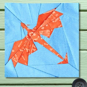 May include: A square fabric block featuring a stylized orange dragon design against a light blue background. The dragon is composed of geometric shapes with a mottled pattern. The background is a patchwork of light blue triangles.