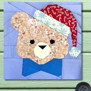 Peut inclure: Un patchwork de tissu représentant un ours en peluche portant un bonnet de Père Noël et un nœud papillon bleu. Le visage de l'ours est fait de tissu floral beige et marron, avec des yeux noirs et un museau blanc. Le bonnet de Noël est rouge avec des flocons de neige blancs et des motifs de cannes en sucre.