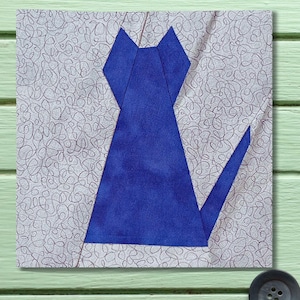 May include: A square fabric block featuring a geometric design of a blue cat silhouette. The cat is centered on a light gray background with a swirling pattern. The block is set against a light green wooden surface.